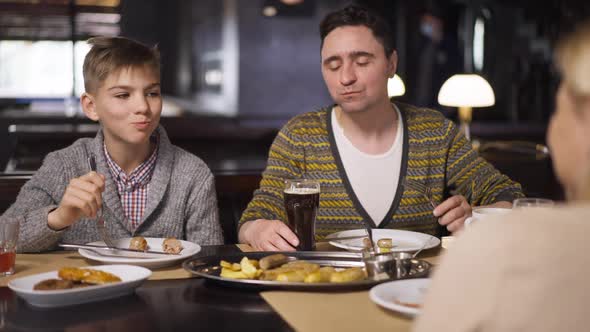 Cheerful Caucasian Father and Son Smiling Eating Tasty Food in Restaurant Sitting at Table with Wife