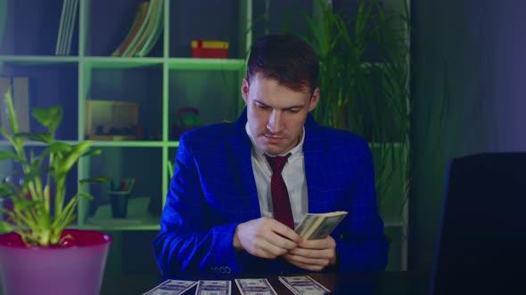 Young Businessman Counts Money Putting Them in Piles on Table in Office