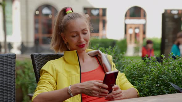 a Blonde in Red and Yellow Clothes is Sitting with a Phone in Hands at a Cafe Table Against the