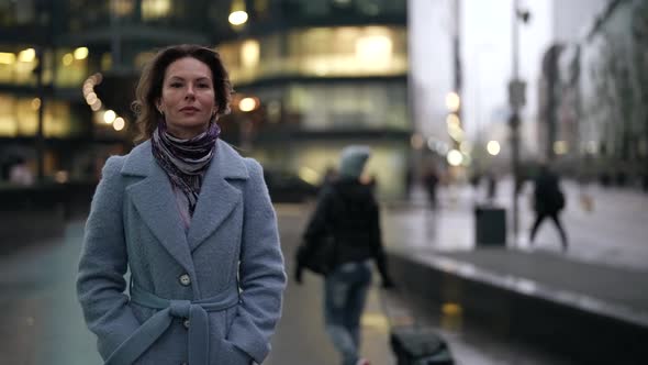 a Woman in a Light Coat and Scarf Has Hands in Pockets and is Walking Along a City Street at Dusk