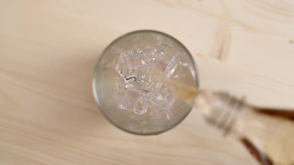 Top View of Cream Soda Drink Being Poured in a Glass with Ice Cubes on Light Brown Wooden Table