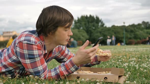 Man Lying on the Grass Eating Pizza and Using a Smartphone in a City Park on Nature