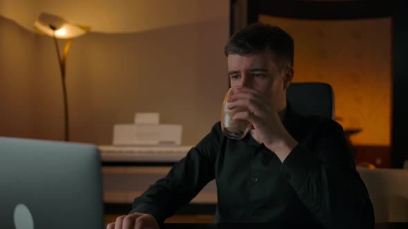 Caucasian Male Drinks Coffee at Home in the Evening Looking at His Laptop