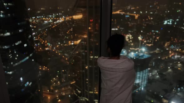 Attractive Woman Wrapped in a Blanket Looks Out the Window at the Lights of the Night City in a