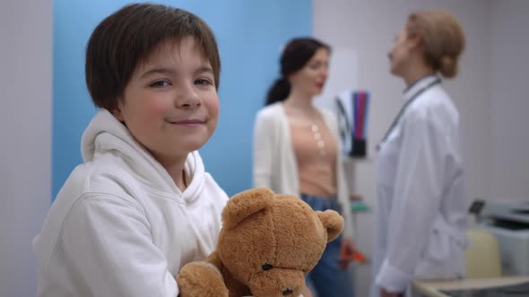 Portrait of Happy Boy Looking at Camera Smiling Hugging Teddy Bear As Blurred Woman and Doctor