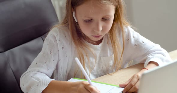 Focused Blonde Schoolgirl Sits at Table at Home, Writes Task in Notebook with Pen, Does Homework