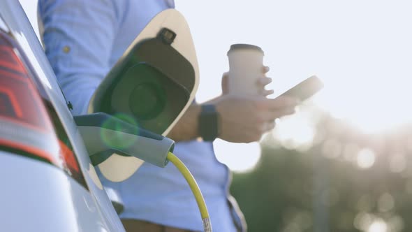 Man Standing Near an Electric Car that is Charging and Making Time Adjustments on Smartphone