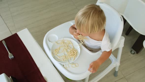 Baby Boy Eats Spaghetti Sitting in a Baby Chair in Restaurant