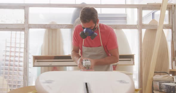 Caucasian male surfboard maker wearing a breathing face mask and shaping a wooden surfboard