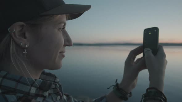 Happy Woman Photographing Sunset over Lake with Phone