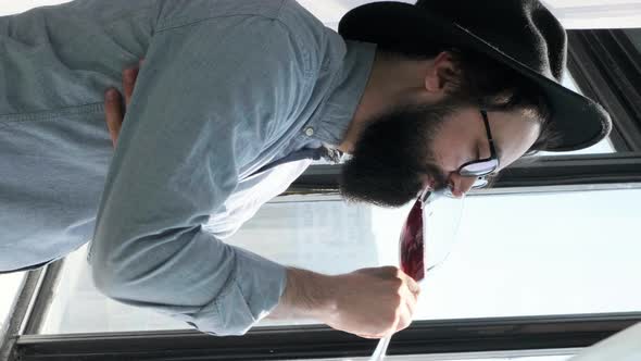 Pleased Bearded Man Drinking Red Wine and Admiring View Form Window at Home