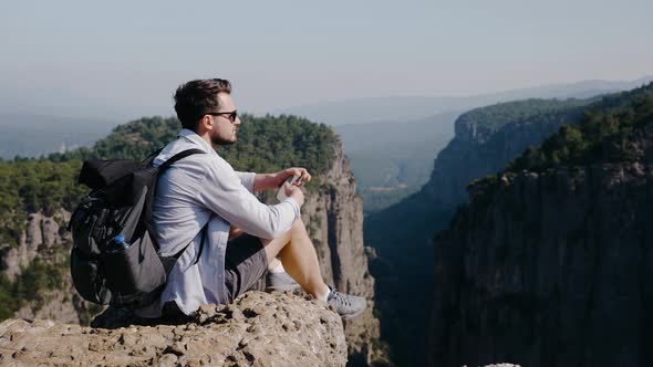 Traveler with Backpack Sits on Cliff and Photographs a Picturesque View