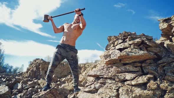 Muscular athlete with hammer on the rocks. Shirtless sportsman breaking stones with hard metal hamme