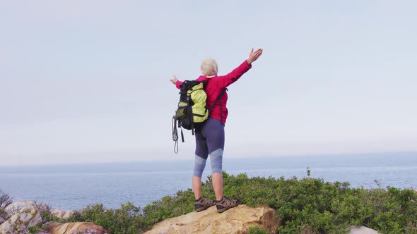 Rear view of senior hiker woman standing with her arms wide open standing on a rock while trekking i