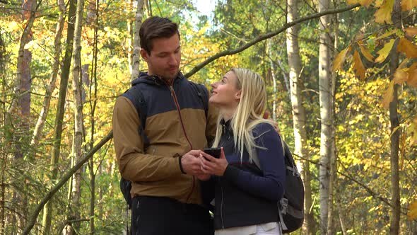 A Hiking Couple Stands in a Forest on a Sunny Day and Looks at a Map on a Smartphone