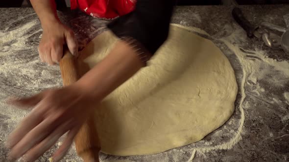 Chef rocks dough with rolling pin on kitchen table against background of flour