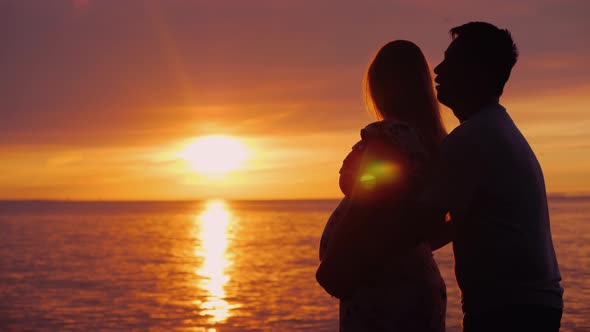 Asian Man Hugs His Pregnant Wife on a Sunset Background Over the Sea