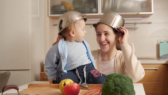 Happy Laughing Mother with Baby Son Playing on Kitchen with Cooking Pans on Heads