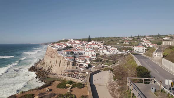 Establishing shot capturing cars visiting Azenhas do Mar town with foamy waves crashing the shore.