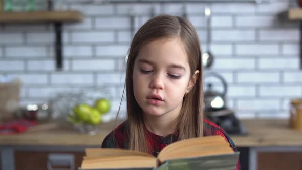 Portrait of a Cute Little Girl in a Plaid Shirt Reading a Book Sitting at the Kitchen Table