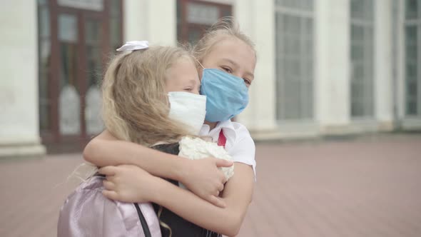 Close-up of Two Schoolgirls in Covid-19 Face Masks Hugging Outdoors. Portrait of Happy Caucasian