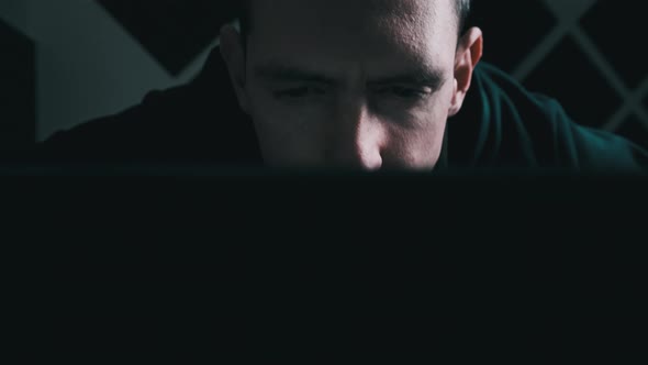 Young Man Looks at a Laptop Monitor Screen in a Dark Room at Home