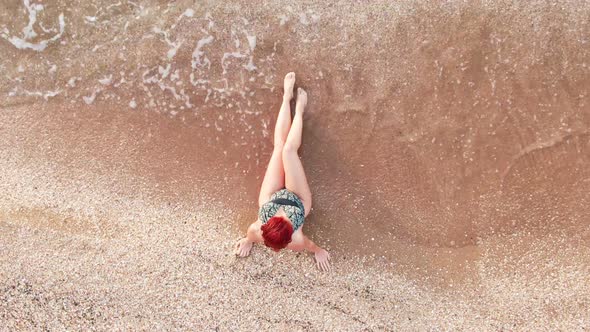 Top View Young Woman in a Swimsuit Sitting on a Sandy Beach Her Legs Caressing the Surf