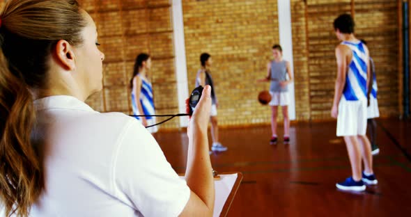 Female coach looking at stopwatch while students playing in basketball court