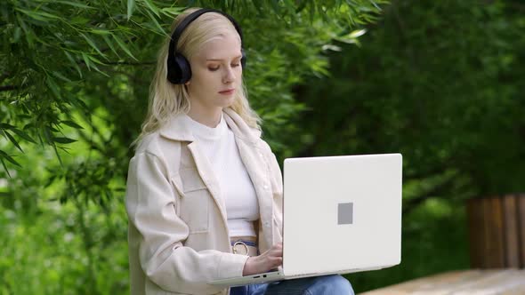 Young Woman in Black Headphones Works Behind a Laptop on a Park Bench