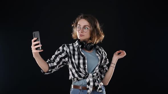 Cute Young Woman in Plaid Shirt and Headphones on Neck Prapering Herself Looking to Her Smartphone