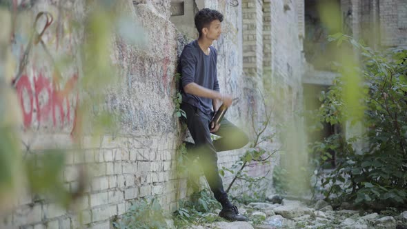 Wide Shot of Frustrated Teenager Standing at Abandoned Ruined Urban Slum and Drinking Beer. Side