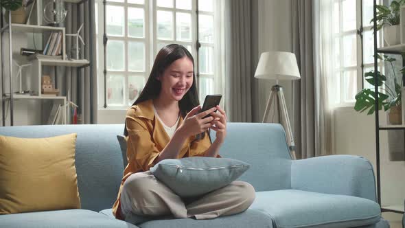 Happy Asian Woman Sitting On Couch In Living Room And Using Smartphone