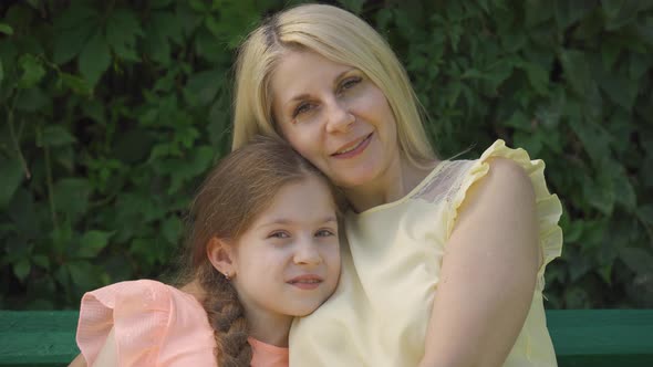 Portrait of Mature Blond Mother in Glasses and Her Little Daughter Looking at Camera While Sitting