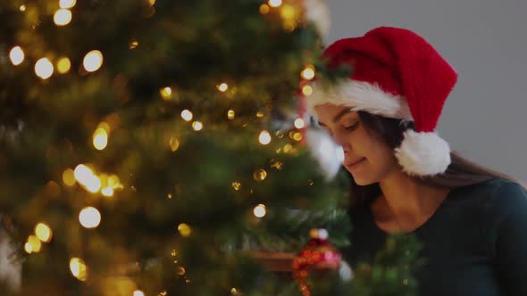 Pretty Young Female in Red Santa Hat Looking Christmas Tree Touching Ornaments