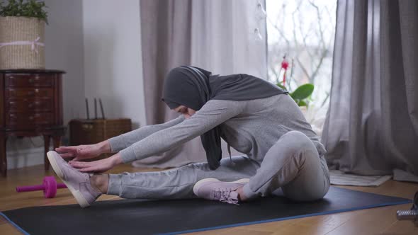 Side View of Slim Muslim Woman Bending Forward on Yoga Mat Trying To Reach Tiptoes with Hands