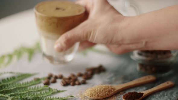 Woman hand with Dalgona coffee with almond milk and brown sugar, coffee beans and green plant