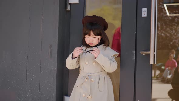 Korean Girl in a Gray Coat and Cap Stands the Street in Spring at the Glass Doors of the Store