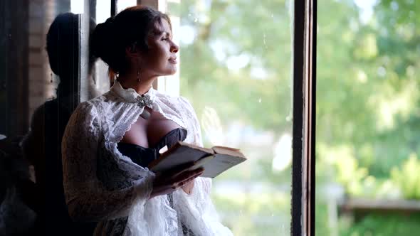 Sexy Woman in Vintage Dress with Open Decollete is Reading Book and Looking at Open Window of