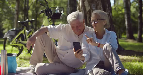 Happy Mature Couple Relaxing in Park on Picnic Using Smartphone Together