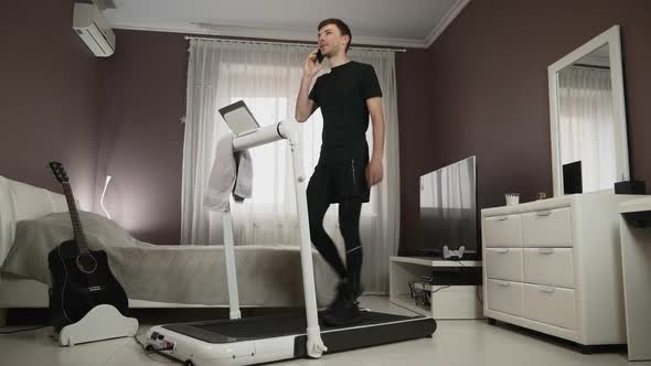 Busy multitasking young businessman is walking on treadmill and talking business on smartphone 