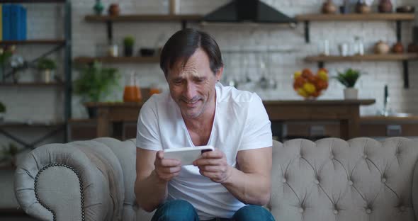 Happy Caucasian Mature Man Gamer Play Video Game Online with Smartphone App Sitting on Sofa at Home