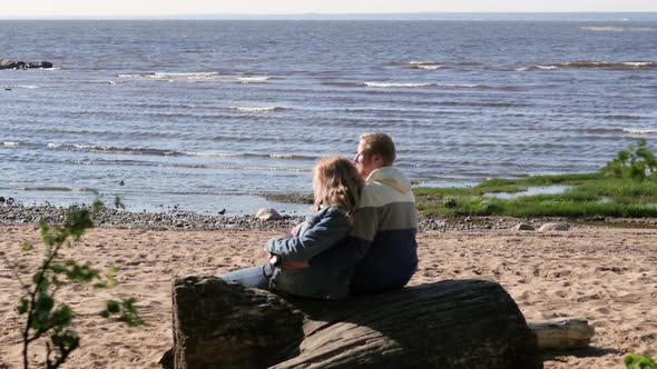 A Pregnant Woman Is Hugging Her Husband Against the Backdrop of the Lake, Sitting on a Log. Young