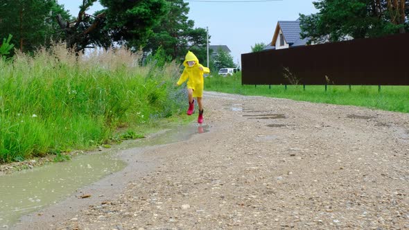 A girl in red rubber boots and a yellow raincoat runs through puddles after a rain in the village. S