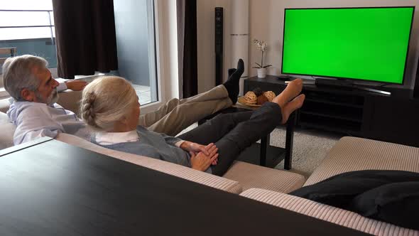 An Elderly Couple Sits in A Living Room in An Apartment and Watches TV with A Green Screen