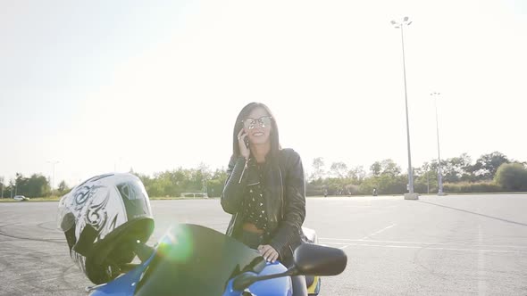 Young Biker Girl with Dark Hair in a Leather Jacket and Sunglasses Sits