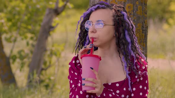 Young Woman in Glasses Red Dress Drinking Refreshing Beverage Sitting Near Tree in Sunny Windy