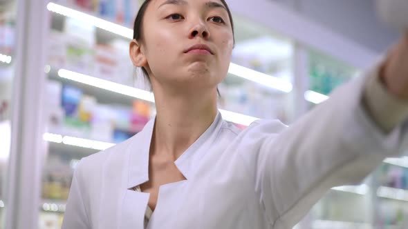 Smart Young Asian Woman Choosing Medication on Shelf in Pharmacy Thinking