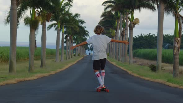 A Teenager with Long Hair Rides a Skateboard Along a Beautiful Road with Palm Trees