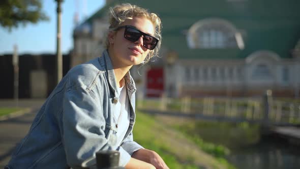 Slow Motion Side View of Charming Blond Girl in Sunglasses and Jacket Leaning on Fence Looking at