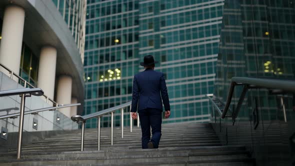 a Stylish Man in a Plaid Suit and Hat Climbs the Stairs Against the Background of a Blurry Modern